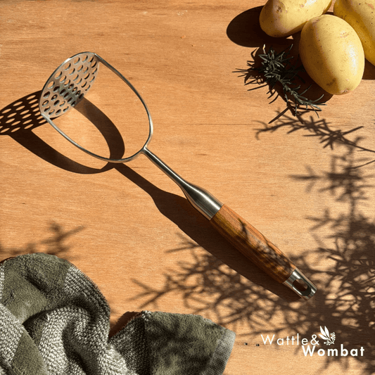 Metal potato masher with wooden handle on a wooden surface with potatoes and a towel.