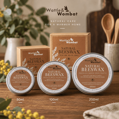 Three Wattle & Wombat Natural Beeswax tins in 50ml, 100ml, and 200ml sizes arranged on a wooden surface with their matching brown cardboard boxes behind them. The scene includes warm home-style props such as a ceramic vase, wooden spoon in a cup, and soft floral decor.